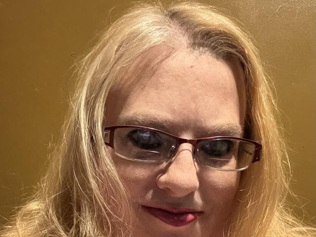 Close-up de Cherish Williams, uma mulher loira de pele clara, usando óculos de aro preto, com um leve sorriso, batom vermelho e cabelo despenteado, contra um fundo marrom simples, mostrando uma tensão sexual sutil..