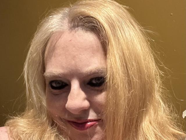 Primo piano di Cherish Williams, una donna bionda con la pelle chiara e occhi intensi, labbra rosse leggermente dischiuse, capelli spettinati, e sfondo scuro.
lei guarda direttamente la fotocamera con uno sguardo provocante.