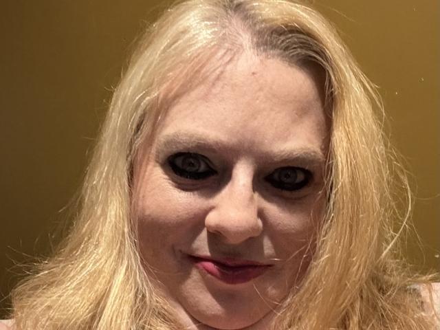 Primo piano di Cherish Williams, una donna bionda con la pelle chiara e gli occhi scuri, che sorride direttamente alla telecamera. I suoi capelli spettinati incorniciano il suo viso, creando una tensione erotica con il suo sguardo diretto..