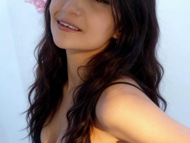 Close-up of AriannaSeete, focusing on her face and upper body. She has long, wavy black hair, light skin, and a slight smile. She's wearing a black top, with sunlight highlighting her face.