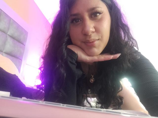 Close-up selfie van LunaDamasco, een mollige vrouw met lang zwart haar, die haar kin op haar hand heeft rusten en recht in de camera kijkt. De camera, met een subtiele glimlach. De paarse achtergrondverlichting accentueert haar gezicht..