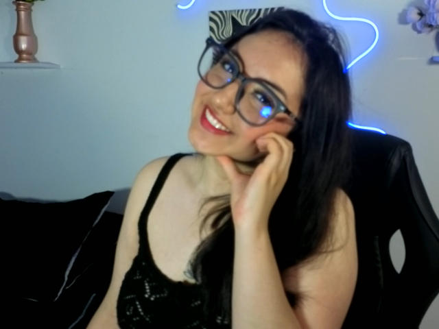 SusanMonroy, con gafas y una blusa negra de encaje, sonriendo de forma seductora, con la mano en la mejilla, luces de neón azules en el fondo, ambiente oscuro. cabello, piel pálida, aura sensual, insinuación de deseo en su mirada, pose relajada..