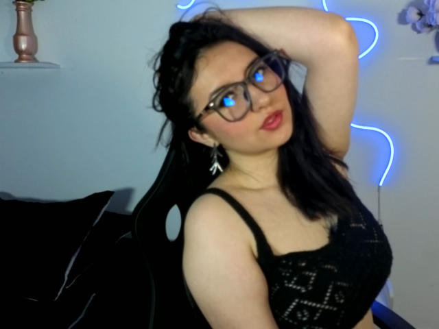 Foto van SusanMonroy, een aantrekkelijke Aziatische vrouw met zwart haar, rode lippenstift en een bril, die een zwarte kanten top draagt, hand in haar, blauwe neonlichten op de achtergrond, intense blik, verleidelijke pose.