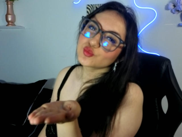 SusanMonroy, a woman with long black hair and blue glowing glasses, pouts her lips while extending her hand forward. She's wearing a black top, sitting against a blue neon light background.