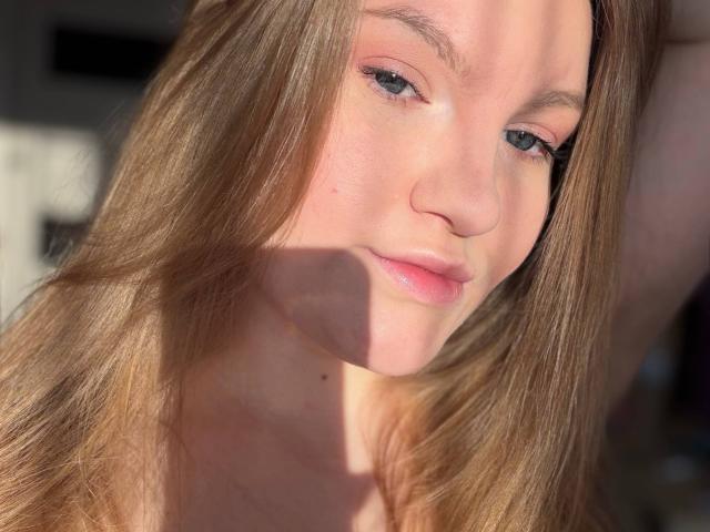 Primer plano de AshleyJonse, la luz del sol iluminando su rostro, cabello castaño largo, ligera sonrisa, sombra en su mejilla, aspecto suave. luz sobre su piel, un ligero indicio de tensión sexual por el contacto visual directo.