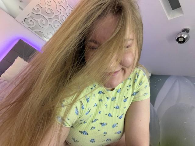 Photo en gros plan d'AshleyJonse, cheveux blonds partiellement cachant son visage, portant un crop top jaune avec des motifs bleus. Légèrement souriant, peau claire, fond violet et blanc, pose suggestive, éclairage naturel..