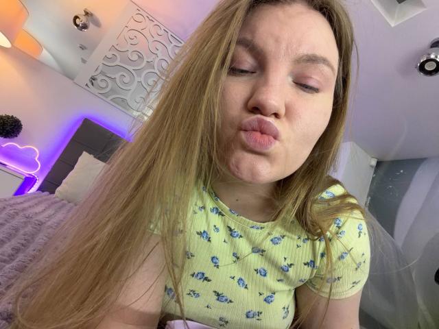 Close-up foto van AshleyJonse met lang bruin haar, volle lippen en gesloten ogen, die een gele blouse draagt met blauwe bloemen. patronen. De achtergrond toont een zwak verlichte kamer met neonlichten. Super schattig..