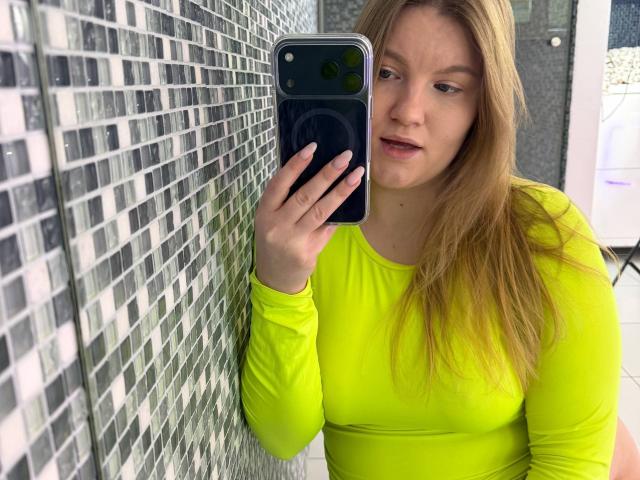 AshleyJonse, with long blonde hair, wearing a tight, bright yellow long-sleeve shirt, holds a black smartphone to her face, leaning against a wall of black and white tiles. Fucking sexy.
