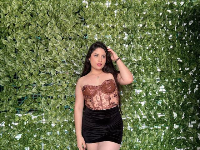 LucianaBlake stands against a lush green leafy wall, wearing a sheer brown lace top revealing her small breasts and a tight black skirt. She touches her hair with a sultry expression.
