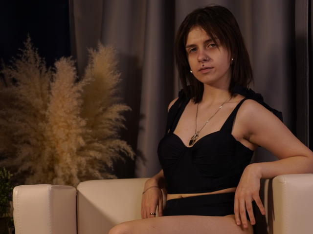 DejaSorley sits in a white armchair, wearing a black crop top and shorts, with a necklace. Dark hair, neutral expression, hands on thighs. Pampas grass in background. Dim lighting.