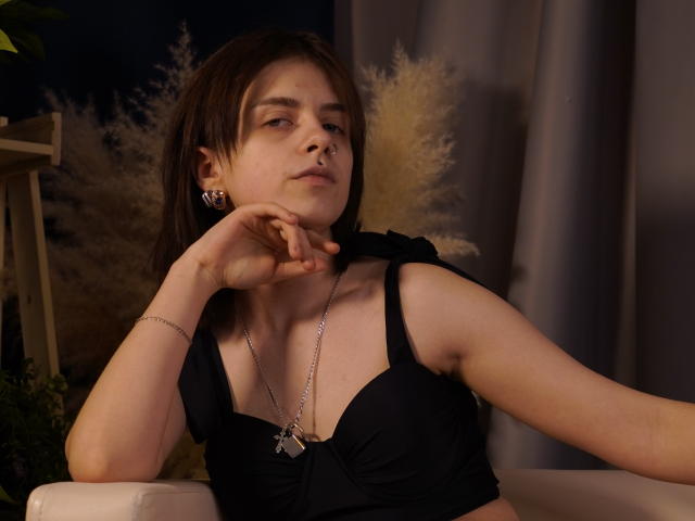 DejaSorley met kort donker haar, een zwarte tanktop, een zilveren ketting en oorbellen, leunt tegen een witte stoel, met haar hand rustend. op het gezicht, zachte belichting, donkere achtergrond, lichtelijk zelfingenomen uitdrukking, sexy uitstraling.