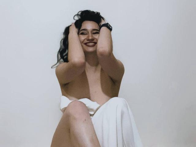 Fotografia di MaryLori, senza top, con un asciugamano bianco che le copre l'area inguinale, mani tra i capelli scuri, sorridente, con un sorriso luminoso. Contesto. Il suo pene è nascosto, ma la tensione sessuale è evidente nel suo sorriso..