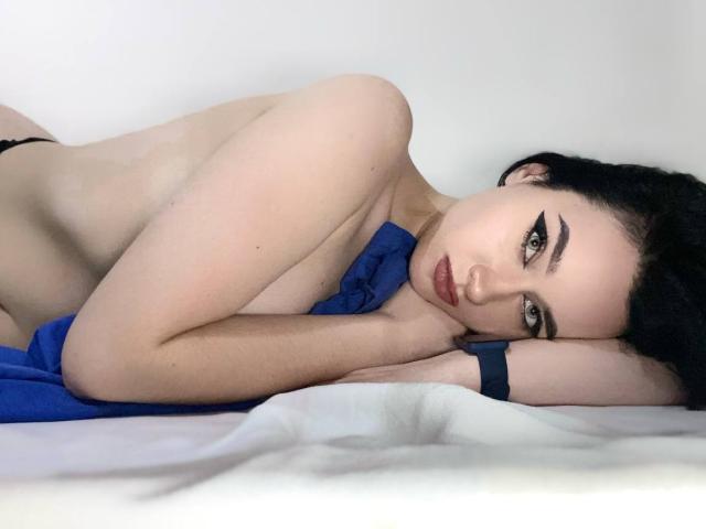 MaryLori liegt auf einem weißen Bett, oben ohne, wobei ein blaues Tuch ihren Brustkorb teilweise bedeckt. Sie blickt verführerisch in die Kamera und trägt ein schwarzes Armband. Dunkles Haar, rote Lippen.