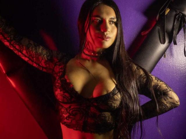 Fotografia de GreizSubKimura com cabelos longos e pretos, vestindo uma blusa preta de renda com decote baixo, que revela o colo, com um fundo roxo. parede. A iluminação em vermelho e preto cria sombras intensas e tensão sexual..