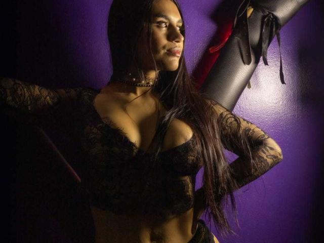 Photographie de GreizSubKimura, une femme à la peau foncée avec de longs cheveux noirs, portant un haut en dentelle noire avec un décolleté plongeant. Collier ras du cou, main sur la hanche, debout devant un mur violet, regard intense, tension sexuelle..