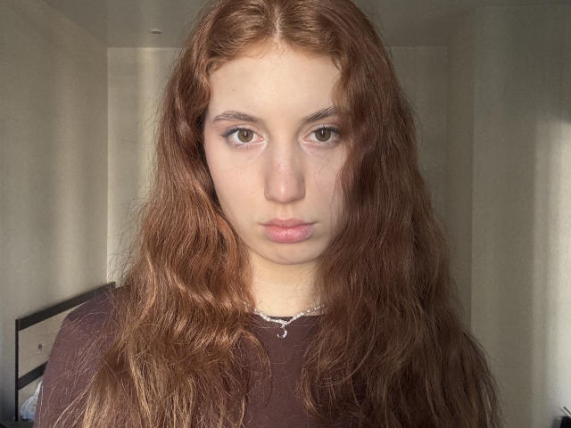 ZeldaEvart, a young woman with long, wavy brown hair, stares directly at the camera with a neutral expression. She wears a black shirt, sunlight highlights her face, intense gaze.