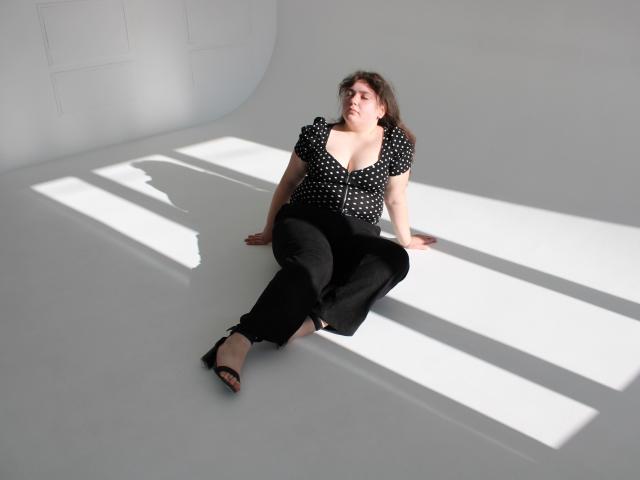 NeonilaLisova zit op een witte vloer, met gespreide benen, een zwart topje met stippen, zwarte broek en zwarte hakken. Het zonlicht creëert scherpe schaduwen. Ze kijkt omhoog, met een ontspannen, enigszins verleidelijke uitdrukking. Ontspannen, aantrekkelijke pose.