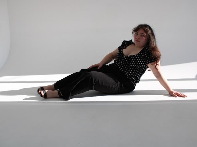 NeonilaLisova est allongée sur un sol blanc éclatant, les jambes tendues, appuyée sur un bras. Elle porte un haut noir à pois, un pantalon noir et des sandales noires. Son expression est détendue, et elle maintient un contact visuel direct..