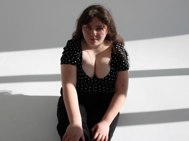 Neonila Lisova beugt sich vor, die Hände auf den Knien, und trägt ein schwarzes, gepunktetes Kleid mit tiefem Ausschnitt, das ihre Brust betont. Das Sonnenlicht wirft Schatten auf ihren geschwungenen Körper. Sie hat dunkles, welliges Haar..
