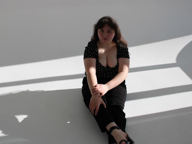 Neonila Lisova est assise par terre, les jambes croisées, vêtue d'un haut noir à pois et d'un pantalon noir. La lumière du soleil projette des ombres nettes sur son corps, mettant en valeur sa poitrine généreuse. Elle regarde en avant.
.