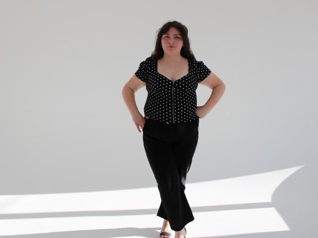 Neonila Lisova se encuentra con confianza frente a un fondo blanco brillante, vestida con un vestido negro con lunares y pantalones negros. manos en las caderas, pelo largo y oscuro, expresión neutra, sombra blanca en el suelo.