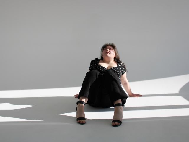 Neonila Lisova, sentada no chão branco com as pernas abertas, vestindo uma blusa preta com bolinhas, calças pretas, saltos altos pretos, cabelos escuros. luz suave, olhar intenso, pose casual, leve sombra no lado esquerdo dela.