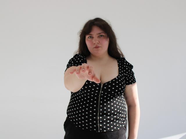 NeonilaLisova, plus-sized woman with long dark hair, wearing a black polka dot top with a zipper, reaching out with her right hand, looking directly at the camera with a serious expression.