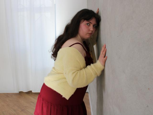 Neonila Lisova, con su largo cabello negro y rizado, apoyada contra una pared blanca con las manos. Vestida con un suéter amarillo y una falda roja, mostrando una pose sensual y provocativa. Luz natural y brillante.