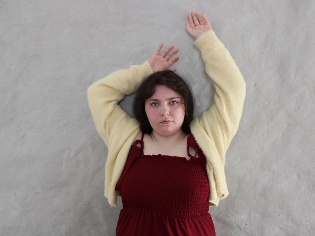 NeonilaLisova lying on a textured gray surface, arms above head, wearing a cream cardigan over a maroon dress, looking straight ahead with a slightly annoyed expression. Hands are relaxed.