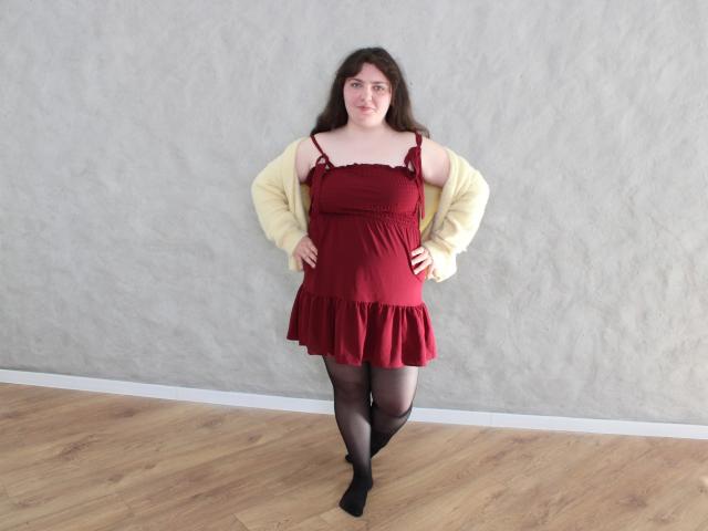 Neonila Lisova steht selbstbewusst da, die Hände in den Hüften, und trägt ein rotes Kleid mit schwarzen Trägern, transparente schwarze Strumpfhosen und gelber Strickpullover.
ihre dunklen, welligen Haare umrahmen ihr Gesicht, und sie blickt direkt in die Kamera..