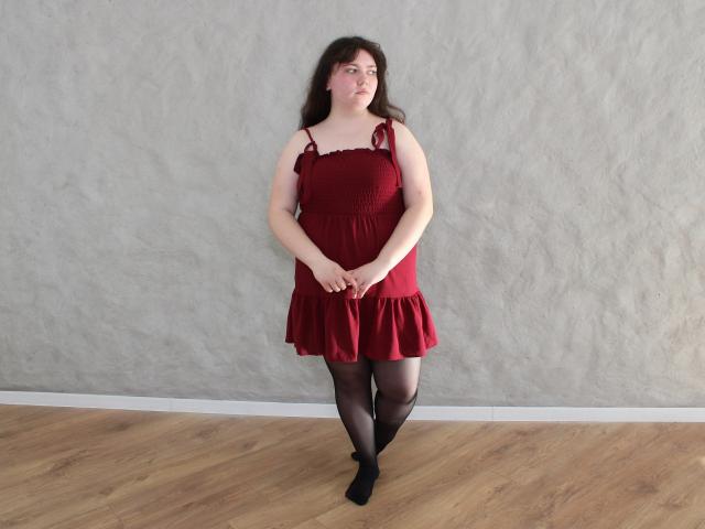 Neonila Lisova, die vor einer strukturierten grauen Wand steht, trägt ein rotes Kleid mit schwarzen Spitzenbändern, schwarze Strumpfhosen und schwarze Schuhe. An die Wand gelehnt, Arme verschränkt, mit einem subtilen, verführerischen Blick..