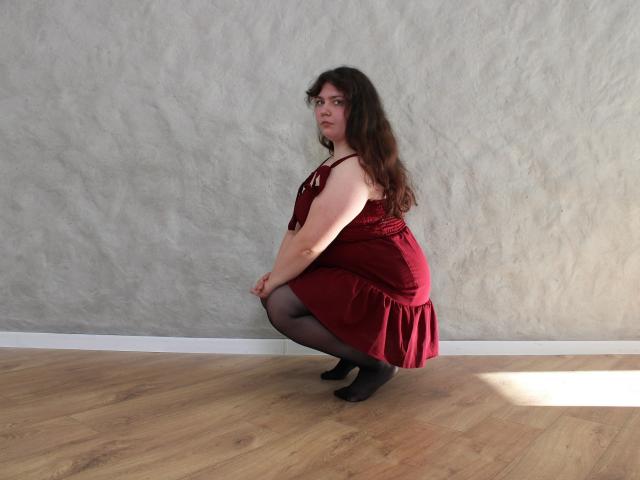 Neonila Lisova, eine wohlgeformte Frau mit langen braunen Haaren, kauert auf einem Holzboden und trägt ein rotes Kleid, schwarze Strumpfhosen und Schwarz schuhe. Graue, strukturierte Wand im Hintergrund, Sonnenlicht auf dem Boden. Verdammt sexy Pose..