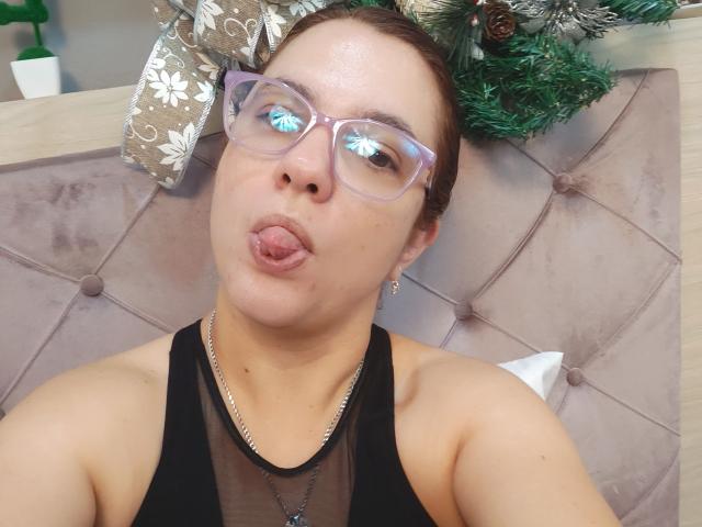 Amelia Wilson, met kort bruin haar, die heldere brillen draagt en een zwarte top van gaas, steekt haar tong uit. Haar tong raakt haar neus. Op de achtergrond is een gestoffeerd hoofdeinde en kerstdecoratie te zien..