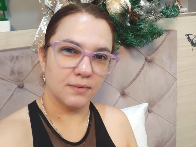 Amelia Wilson heeft bleke huid, bruin haar in een staart, en draagt paarse brillen. Ze draagt een zwarte top met een zilveren ketting. Op de achtergrond staat een kerstboom..