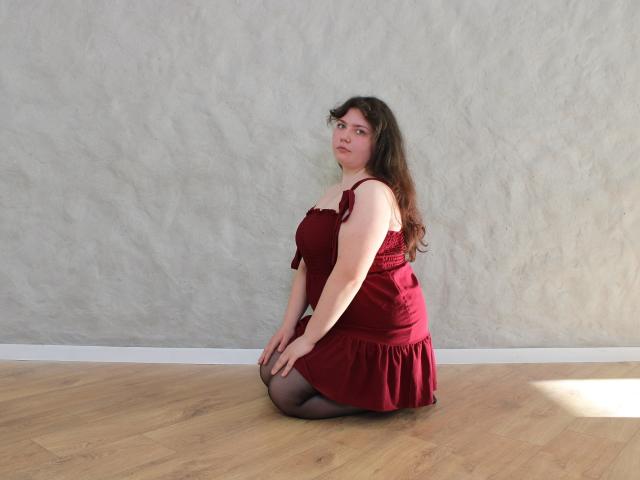 Neonila Lisova kniet auf dem Holzboden und trägt ein rotes Kleid und schwarze Strumpfhosen. Sie hat langes, dunkles Haar, helle Haut und blickt mit ernstem Gesichtsausdruck über ihre Schulter. Sonnenlicht wirft Schatten..