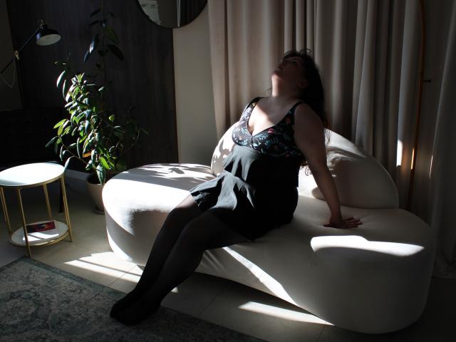 Neonila Lisova è seduta su un divano bianco e curvo in una stanza poco illuminata, illuminata dalla luce del sole. Indossa un abito nero, ricoperto di paillettes, con la schiena arcuata, gli occhi chiusi e le gambe leggermente divaricate. Tavolo con piante e decorazioni dorate nelle vicinanze..