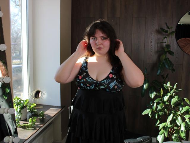 Neonila Lisova, von kräftiger Statur, mit heller Haut und dunklem Haar, steht drinnen und richtet ihre Haare. Sie trägt ein schwarzes Kleid mit tiefem Ausschnitt und bunten Mustern, das Sonnenlicht betont ihre Brust. Pflanzen in Töpfen umgeben sie..