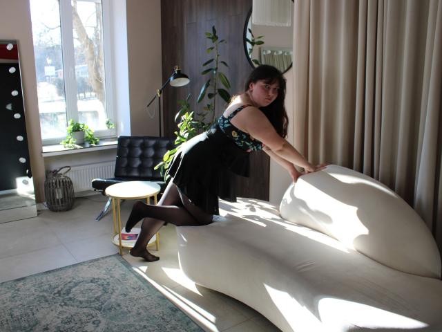 Neonila Lisova lehnt sich auf ein cremefarbenes Sofa, während Sonnenlicht durch ein Fenster scheint. Sie trägt ein schwarzes Kleid und schwarze Strumpfhosen. Schatten fallen über ihren Körper, Pflanze im Hintergrund, Vorhänge rechts..