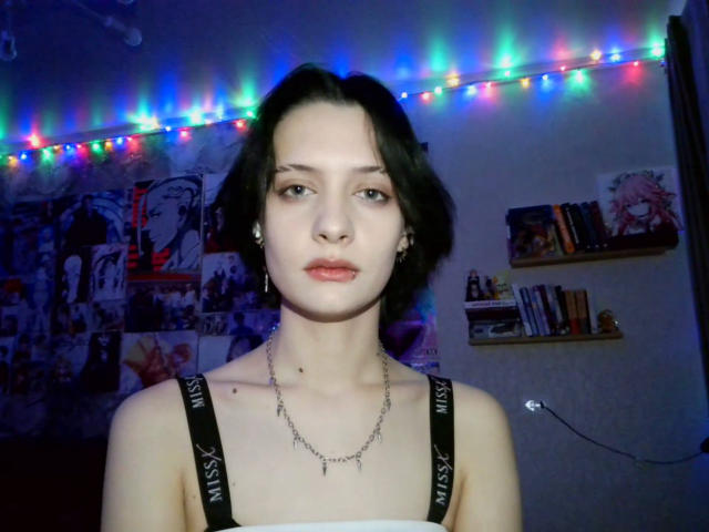 Photograph of MelyndaMarron, front-facing, pale skin, black hair, neutral expression, wearing black strap top, chain necklace, illuminated by colorful string lights, posters on wall, shelves with books. Fucking sexy vibe.
