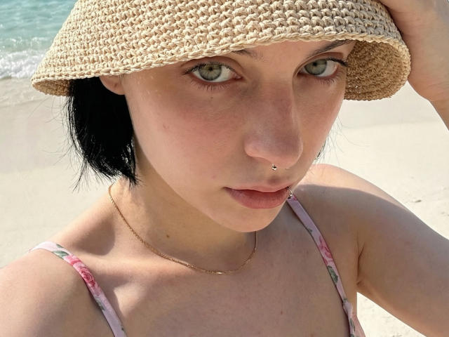 Close-up photo of MelyndaMarron, pale-skinned with black hair, wearing a straw hat and floral bikini top, piercing nose, blue eyes, holding hat, beach background, sunlight highlighting face, sexual tension.