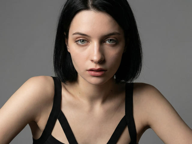Close-up photograph of MelyndaMarron, fair-skinned with black shoulder-length hair, wearing a black strappy top, direct gaze, slightly parted lips, light shadows, grey background, intense and sexual tension.