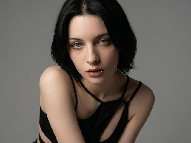 Photographie en gros plan de MelyndaMarron, avec une peau pâle et une coupe de cheveux courte noire, regardant intensément avec de grands yeux bleus. Portant un haut noir à bretelles, penchée en avant, créant une tension sexuelle intense..
