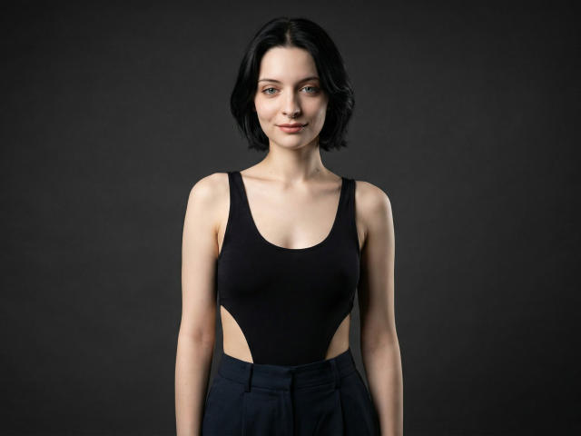 Photograph of MelyndaMarron with short black hair, wearing a black tank top with side cutouts, standing against a dark background. She has a confident expression, minimal makeup, and visible cleavage.