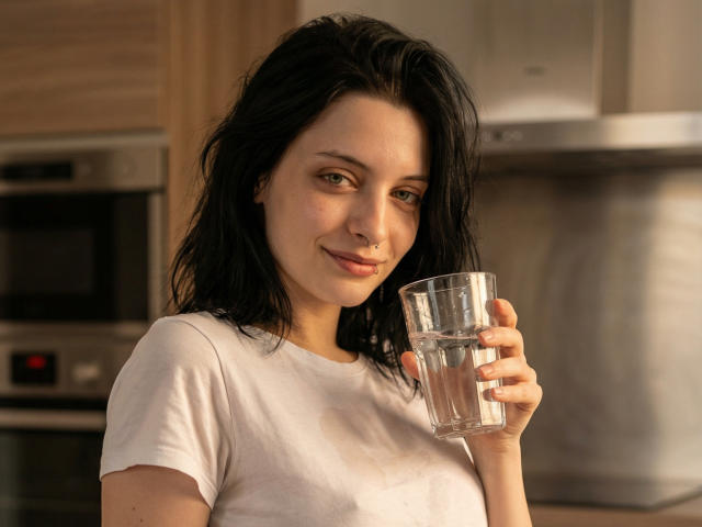 MelyndaMarron, com cabelos pretos desalinhados, segura um copo de vidro transparente com água em uma cozinha moderna. Ela veste uma camiseta branca, sorrindo levemente, com uma sugestão sutil de sensualidade. A luz suave realça seu rosto..