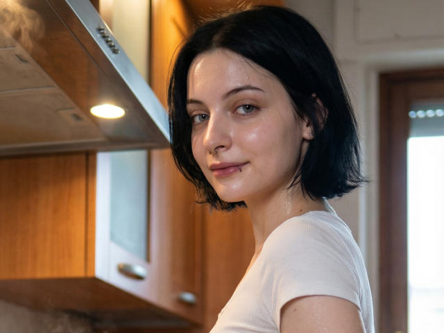Photograph of MelyndaMarron with short black hair, pale skin, and a nose ring, wearing a white t-shirt, looking over her shoulder, standing in a wooden kitchen, light in background.