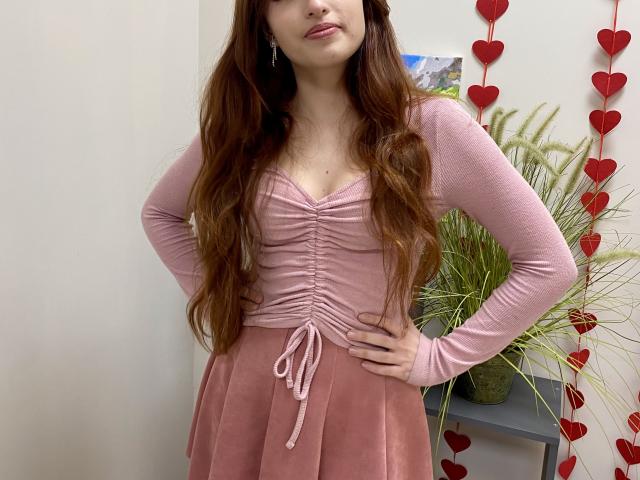 Rioria with long brown hair, wearing a pink ribbed top and matching skirt, stands with hands on hips, red heart decorations hanging in the background, cock implied by pose.