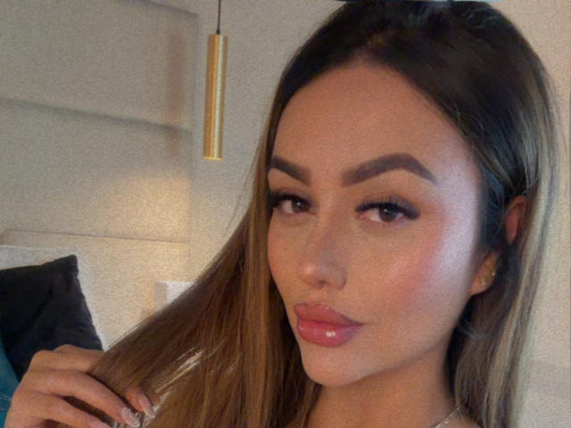 Gros plan de MissAry, une femme séduisante avec de longs cheveux bruns, des sourcils épais et des lèvres roses brillantes. Elle tient ses cheveux avec une main, et regarde directement la caméra, avec une expression séduisante..