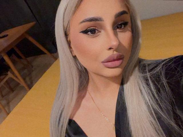 Primo piano di CurvyVanessa con lunghi capelli lisci, biondo platino, sopracciglia folte, eyeliner scuro e rossetto rosa. Indossa una maglietta nera, guarda direttamente la fotocamera con un'espressione seria..