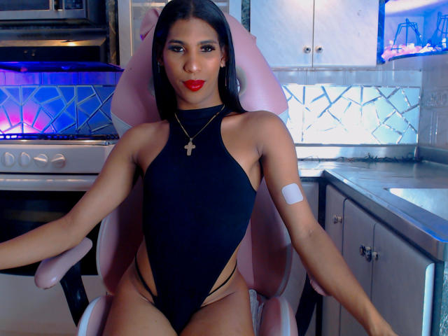 MirandaPresly, seated in a pink chair, wears a black, high-cut leotard with side cutouts, red lipstick, and a cross necklace. A white bandage on her upper arm. Kitchen background.