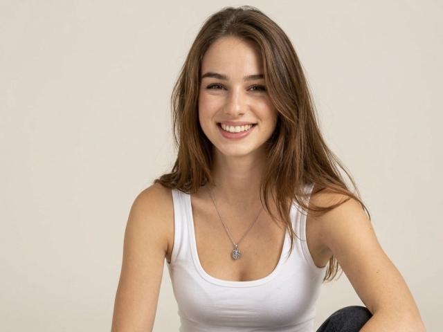 StellaNoir, uma jovem com longos cabelos castanhos, sorri diretamente para a câmera. Ela veste uma camiseta branca, que revela uma silhueta esguia e seios pequenos. Fundo cinza claro.